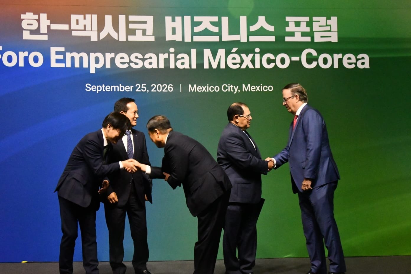 Participantes se saludan en el escenario del Foro Empresarial México-Corea, frente al nombre del encuentro.