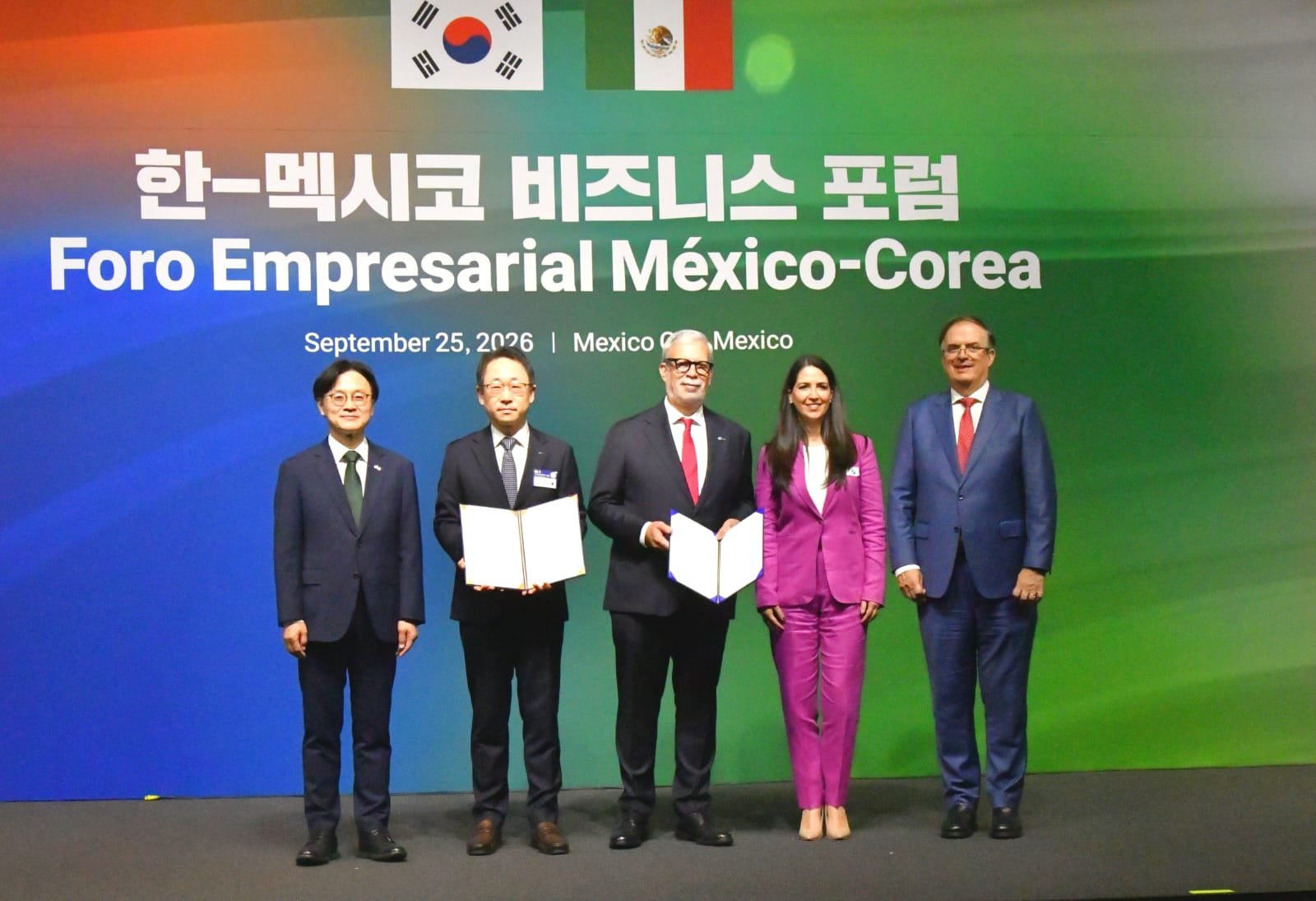 Cinco participantes posan frente al escenario del Foro Empresarial México-Corea; dos de ellos muestran documentos abiertos.